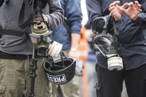 image or two male reporters holding gas masks, a tripod and a helmet with a “PRESS" imprint.