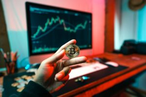 A person holding a physical Bitcoin token in front of a computer screen displaying a stock market line chart.