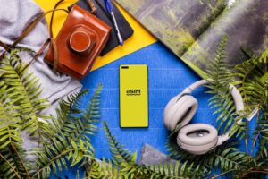 Top-down view of a yellow smartphone with an eSIM logo on a blue surface surrounded by travel gear and ferns.